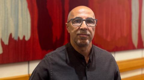 A bald, middle aged man wearing a black shirt and glasses. There is a large colourful art work on the wall behind him. 