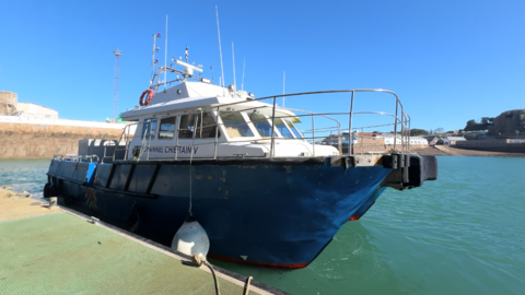 A blue and white vessel named Channel Chieftain V moored at Victoria Harbour. The boat has a sturdy metal rail around the deck, various antennas and equipment on the roof, and a white fender hanging from the side. It sits in calm turquoise water, with a harbour wall and nearby buildings visible in the background under clear blue sky.
