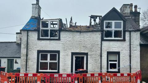 A house in New Abbey which has been hit by fire. The roof has caved in and there are red and white barriers, and police tape, surrounding the building. 