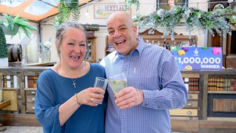 Caroline (left) and Terry Gillings celebrating their win with a drink. Caroline has grey hair, tied back, and is wearing a blue top. Her husband is bald and wearing a blue checked shirt. Behind them is a sign that reads "£1,000,000 winner".