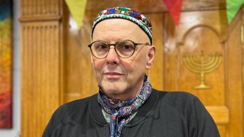 Rabbi René Pfertzel is stood in front of a wooden case and is wearing a neck tie and a yarmulkes as he stands in the synagogue.