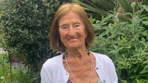 A woman wearing a white cardigan. She is smiling and looking directly at the camera. There are a number of green plants behind her. 