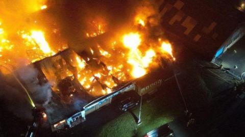 An aerial photo of a blaze at a strip of commercial buildings - the roof has been mostly burnt away showing fierce flames underneath.
