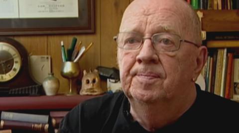 Ian McNicol in 2007 wearing a black tshirt and silver rimmed glasses. In the background there is a bookshelf, piles of books and a clock.