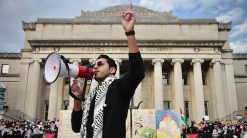 Mohsen Mahdawi with a megaphone raises a finger above his head with a keffiyeh around his neck