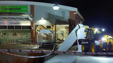 It is dark outside and a Co-op building has been broken into. The front right of the building has had the shutters ripped off and is completely open. A JCB telehandler has been left in front. On the front of the JCB is large metal sheets - which look to be the shutters from the front of the store. Police tape has been used to cordon the scene. 