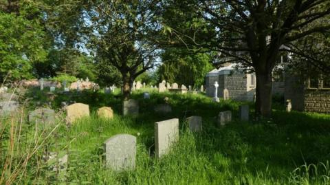 Large stone graves surrounded by long grass in a walled off area to the left of a church. There are multiple large trees dotted around the burial site.