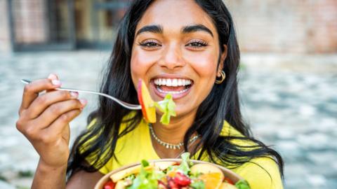 A young woman smiles at the camera, she's holding a fork full of salad in one hand, and in the other she holds the bowl of salad.