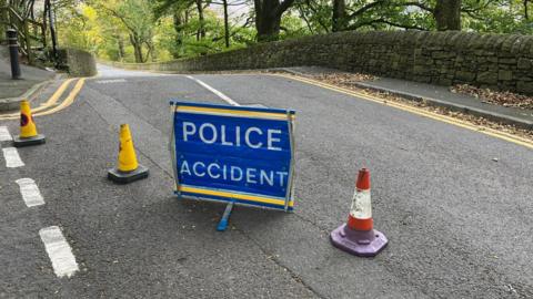 A blue police sign bearing the words 'police accident' beside some cones on a rural road with trees in the background. 