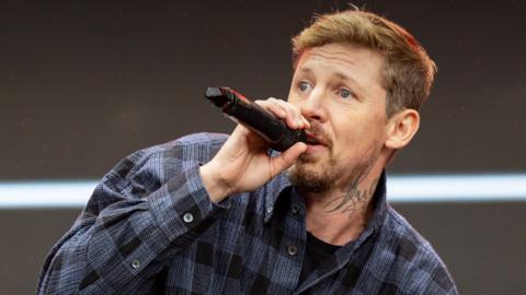 Professor Green wearing a blue and black checkered shirt and holding a microphone.