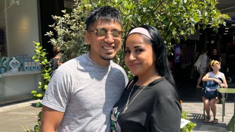 A man and a woman stand together in a sunny plaza. He wears a grey T-shirt and sunglasses. She wears a black T-shirt, necklace and headband