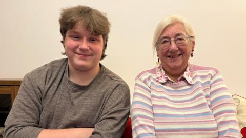 There's a lady sat to the right on the sofa with a pink, white and blue jumper on. On the left there's a younger chap smiling with a grey jumper on. They are both smiling.