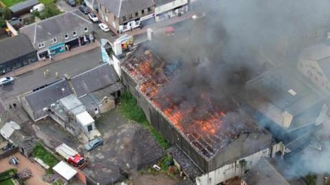 An aerial shot of a lareg fire at a building in Lochgelly. The fire can be seen in the building and is orange. A large plume of dark grey smoke is coming off it.