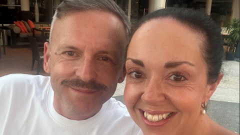 Laura and Paul Webster, a man with shirt grey hair and a mustache is wearing a white tshirt and looking at the camera with a slight smile, a woman next to him has dark brown hair pulled back and pearl earrings and looking at the camera with a wide smile