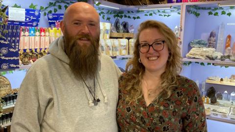 A man with a beard and bald head smiles toward the camera, he is wearing a great hooded top , standing next to him is a woman with blond hair and wearng glasses, she is smiling toward the camera and is wearing a green dress with a brown floral pattern. Behind them are crystals and minerals on display on shelving units.