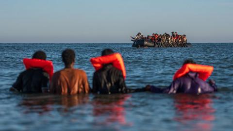 Four people stand in shallow water with life vests on looking out towards a small boat on the horizon in open water in Gravelines, France in August.
