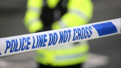 A stock image showing blue and white police cordon tape stretching across the frae, with a police officer wearing a yellow hi-vis coat and black vest standing in the blurred background.