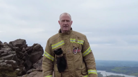 A man with a bald head in a brown firefighter over-suit standing on rocks over a valley below