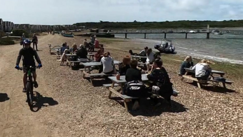 Google Street View of picnic benches on a beach on Mudeford sandbank. It is a sunny day and the benches are filled with people. Two cyclists are passing on the path between the beach and a long row of beach huts.