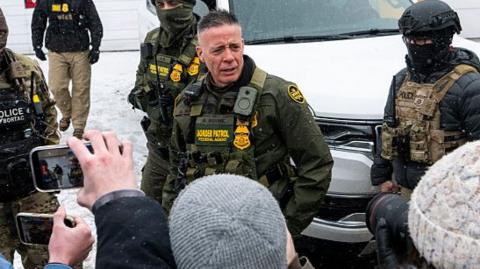 Border Patrol Commander Gregory Bovino, wearing a green and yellow uniform, is confronted by community members on a snow-covered street in Minnesota