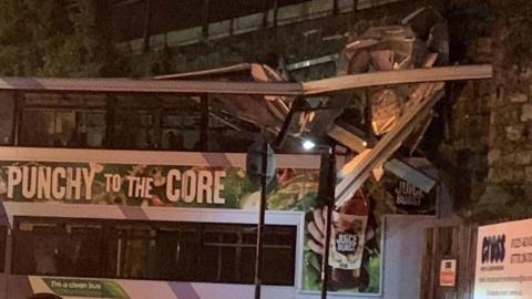 A double decker bus that has crashed into a railway bridge, with a police cordon around the road.