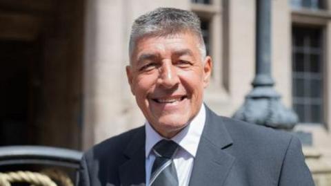 Chris Large is smiling at the camera, he's wearing a suit and tie and standing outside the county buildings on a sunny day. 