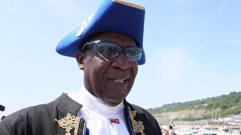 Ed Christopher is wearing traditional town crier uniform in black and blue, along with sunglasses.