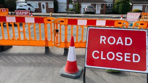 Orange barriers and cones at the side of road resurfacing works on a housing estate. A large red Road Closed sign is on the right.