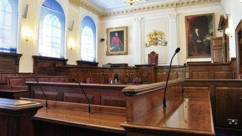 Inside the States of Guernsey chamber. Rows of seating with microphones and an upper row of seats at the back of the room under a crest and two portraits on the wall.