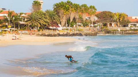 A man surfing near the coastline of Santa Maria, Sal Island, Cape Verde. The water is a pale blue and he is riding a small wave. There are palm trees and white buildings with terracotta-coloured roofs in the background.