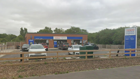 A small Tesco Express shop at the side of a road. A small car park sits in front of the shop with some vehicles parked up. A cash machine can be seen at the left hand side of the shop.