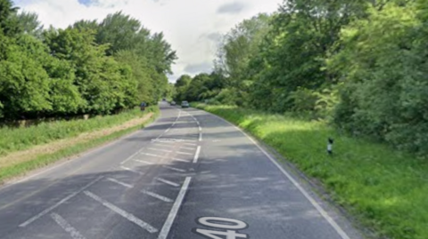 Google street view of the A40 with a car in the distance. There are lots of trees and long grass near the road.