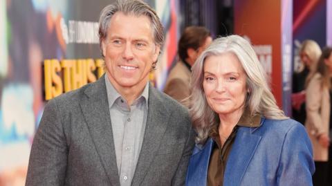 John and Melanie Bishop attending the screening of Is This Thing On? in London. John, with grey hair, is wearing a grey suit and lighter grey shirt. Melanie, with longer grey hair, is wearing a blue jacket and brown shirt. They are both smiling.