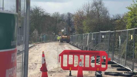 The photo is framed by metal construction fencing on either side. Down the centre we can see a road with its surface stripped away. Cones and red barriers are scattered about, and yellow construction vehicles can be seen in the distance, as well as workers in high vis clothing