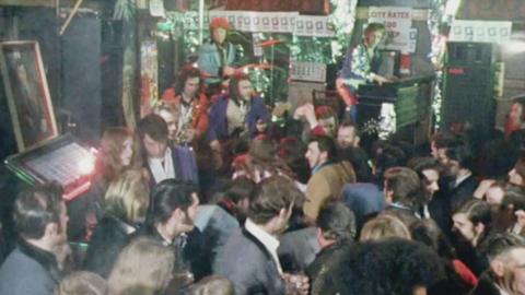 Crowd dancing in pub, with band playing on stage. 