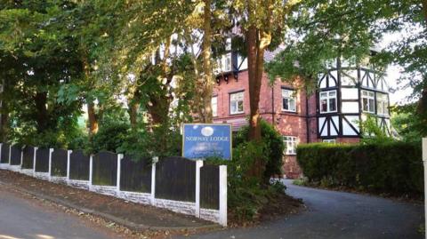 A tree and hedge lined drive way leading to a brick building with Victorian style white fascia lined with black wooden beams. A blue sign marks the entrance with 'Norway Lodge' in white letters. 
