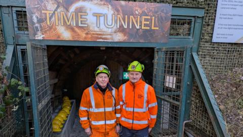 Two men wearing luminous orange jackets and helmets stand beneath a sign reading 'Time tunnel'. 