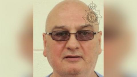 Police custody photograph of a man with a round face and bald head. He is wearing dark rectangular glasses and is staring into the camera.