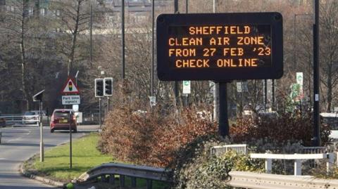 An image of a large electronic sign  which is on a grass verge down the middle of a main road.  The sign reads 'SHEFFIELD CLEAN AIR ZONE FROM 27 FEB '23 CHECK ONLINE'