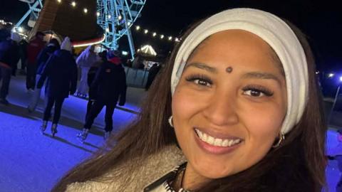 Joyce Leon Velasquez smiles at the camera on an ice rink at Christmas time. She has a white headband on and black coat. People can be seen ice skating behind her. There is also a large Ferris wheel that is lit up.