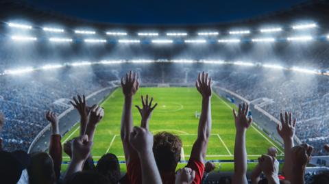 Stock image shows a high angle shot of fans cheering for a team in a big stadium during a football match. The team is playing on the pitch which crowds of fans can be seen celebrating, the image shows a wide angle of a sold out arena.