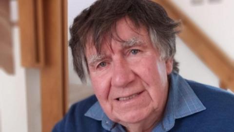 An elderly man with dark hair and grey sideburns with large eyebrows and a blue checked shirt under a blue jumper