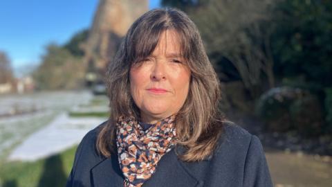 Rachel has bark brown hair. She is wearing pink lipstick and a navy blue coat, with a navy, orange and white scarf. In the background there is snow on the trees and grass.