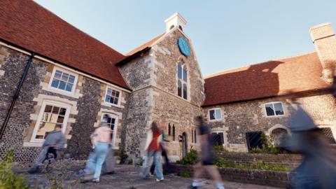 A stone building with a red tiled roof. A line of people are walking in front of it, but their motion is blurred by the camera.