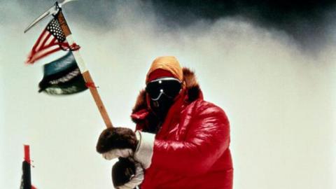 Mountain climber Jim Whittaker wears a red snow jacket, gloves and goggles as he holds a stick affixed with a US flag as he summits Mount Everest in 1963.