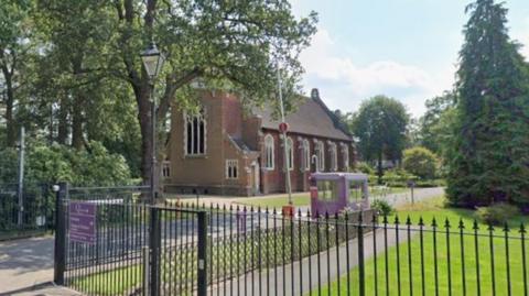 The outside of a school where its name can be seen on a purple plaque. A chapel is in the school grounds.