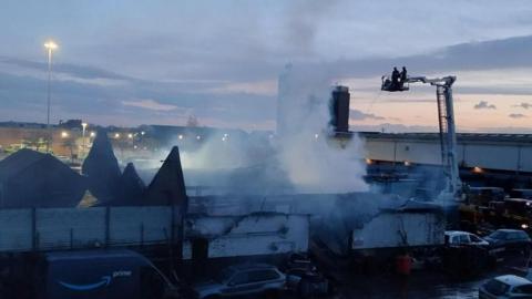 A building badly damaged and smoking after a fire. Firefighters can be seen in a raised platform spraying water onto the structure. Burned debris and several parked vehicles surround the scene. 