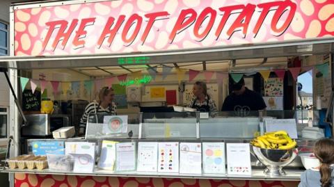 A picture of the spud truck. There is colouring sheets on the front of the truck. There is 3 people stood in the truck.