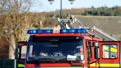 The top half of a red fire engine. The vehicle is red with his visibility stripes. There are blue lights on the top along with ladders. Doors on both sides are open. The front of the vehicle is empty. 