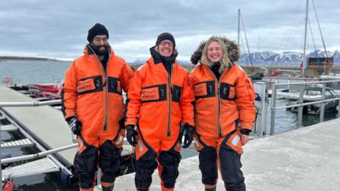 Three researchers from the University of Leicester on their arctic voyage wearing orange overalls. Stood in front of a concrete jetty. Snow-capped mountains are in the background.